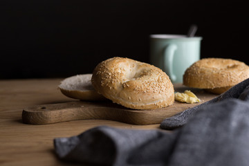 Fresh bagels with sesame and butter lying on a wooden board surrounded by cocoa. Dark background....