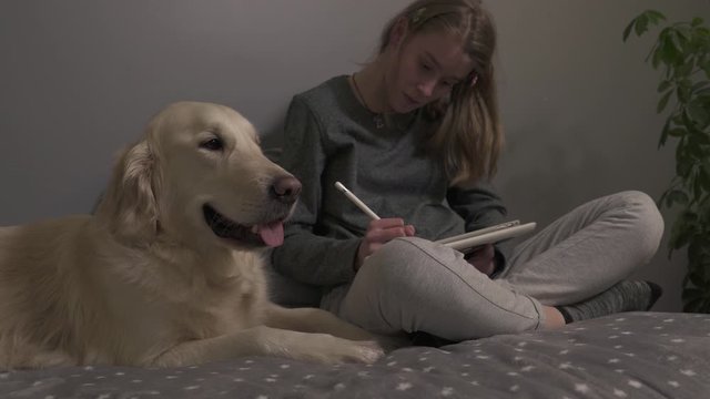 Girl sitting on a bed in an room drawing with her dog.