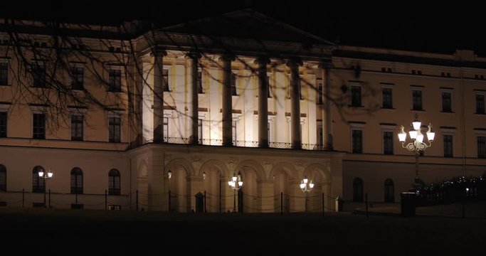 Slow motion wide 4K shot with parallax motion of branches from tree in front of illuminated front entrance pillars of the Norwegian Royal Palace on top of Karl Johan street, at night in Oslo Norway.