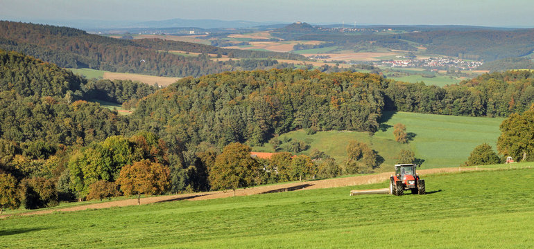 panorama landscape in the Odenwald Germany with forest and meadows, a tactor that mows a meadow. In the background you can see a village and in the distance a castle on a mountain and wind turbines