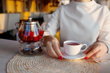 Glass teapot with  tea at cafe. Woman hands with tea cup on background