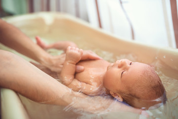 Small cute big-eyed beautiful baby bathes in warm water in the hands of a caring mother. The...