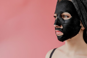 Young woman apply black cosmetic fabric facial mask on pink background. Face peeling mask with charcoal, spa beauty treatment, skincare, cosmetology. Close up