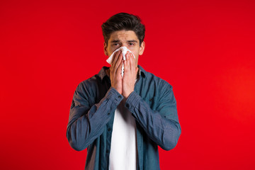 Young man sneezes into tissue. Isolated guy is sick, has a cold or allergic reaction. Coronavirus, epidemic 2020, illness concept. Red background - obrazy, fototapety, plakaty