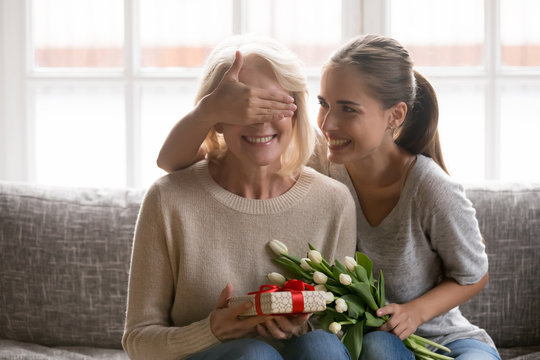Joyful adult millennial daughter covering happy mommys eyes.