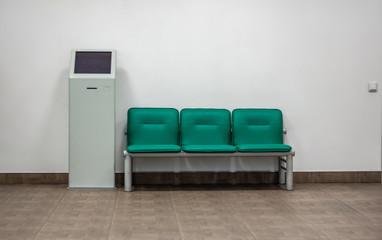 Front view of a row of modern green chairs and terminal against white wall in office waiting room; minimal style interior; waiting, self service and seating area