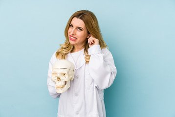 Young doctor woman holding a skull isolated covering ears with hands.