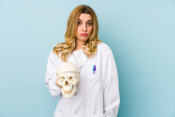 Young doctor woman holding a skull isolated shrugs shoulders and open eyes confused.