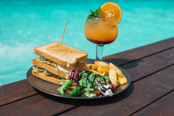 Delicious sandwich  with fries and salad served with cold cocktail next to the pool.