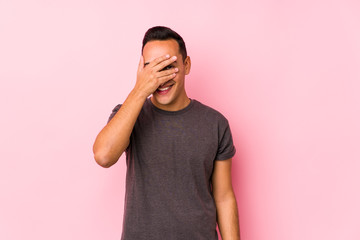 Yooung latin man posing in a pink backgroundblink at the camera through fingers, embarrassed covering face.