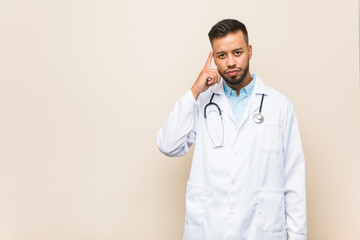 Young south-asian doctor man pointing temple with finger, thinking, focused on a task.