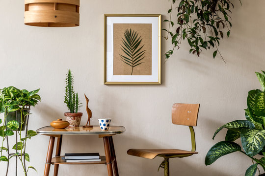 Retro interior design of living room with stylish vintage chair and table, plants, cacti, personal accessories and gold mock up poster frame on the beige wall. Elegant home decor. Template. 