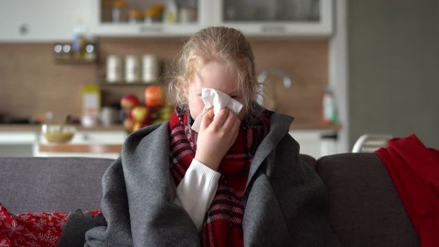 cold caucasian girl at home. portrait of a sick child in a scarf and plaid on the sofa in the apartment, the schoolgirl blows snot into a napkin