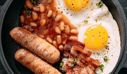 English breakfast on a dark table. fried eggs with bacon canned beans and sausage. near coffee, orange juice and pancakes with butter and honey