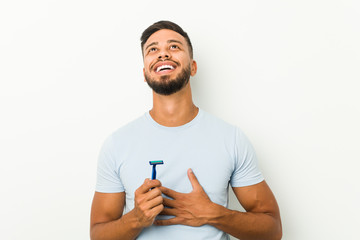 Young south-asian man holding a razor blade laughs out loudly keeping hand on chest.