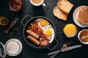 English breakfast on a dark table. fried eggs with bacon canned beans and sausage. near coffee, orange juice and pancakes with butter and honey