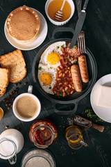 English breakfast on a dark table. fried eggs with bacon canned beans and sausage. near coffee, orange juice and pancakes with butter and honey