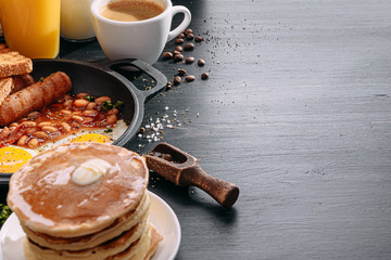 English breakfast on a dark table. fried eggs with bacon canned beans and sausage. near coffee, orange juice and pancakes with butter and honey