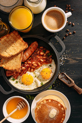 English breakfast on a dark table. fried eggs with bacon canned beans and sausage. near coffee, orange juice and pancakes with butter and honey