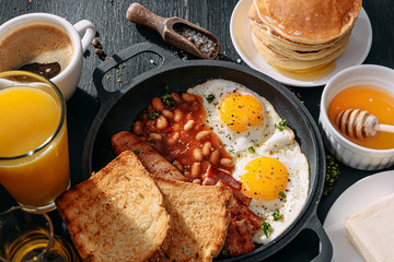 English breakfast on a dark table. fried eggs with bacon canned beans and sausage. near coffee, orange juice and pancakes with butter and honey
