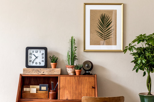 Stylish retro composition of home office interior with vintage wooden cabinet, chair, plants, black clock, cacti, lamp and elegant accessories. Gold mock up poster frame. Retro home decor Template