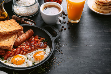 English breakfast on a dark table. fried eggs with bacon canned beans and sausage. near coffee, orange juice and pancakes with butter and honey