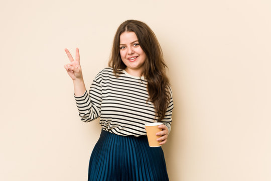 Young curvy woman holding a coffee joyful and carefree showing a peace symbol with fingers.