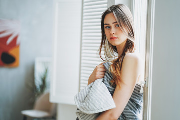 Young beautiful long brown-haired hair girl in blanket near window at bright modern interior, morning routine