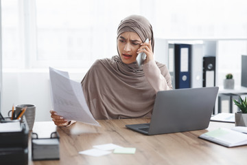 Stressed muslim businesswoman checking documentation and talking on cellphone in office