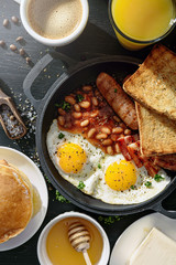 English breakfast on a dark table. fried eggs with bacon canned beans and sausage. near coffee, orange juice and pancakes with butter and honey