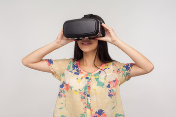 Happy young woman wearing VR glasses and watching virtual world with positive calm facial expression, playing video game, using simulator for education. indoor studio shot isolated on gray background