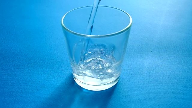 Clear sparkling water pour into a transparent glass on a blue background. Slowmotion.