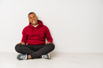 Young latin man sitting on the floor isolated funny and friendly sticking out tongue.
