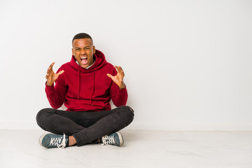 Young latin man sitting on the floor isolated screaming with rage.