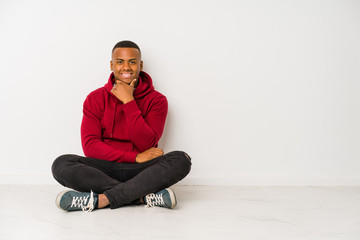 Young latin man sitting on the floor isolated smiling happy and confident, touching chin with hand.