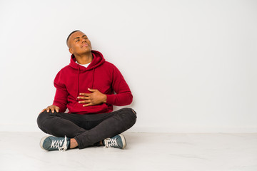 Young latin man sitting on the floor isolated touches tummy, smiles gently, eating and satisfaction concept.