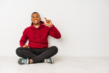 Young latin man sitting on the floor isolated holding something little with forefingers, smiling and confident.