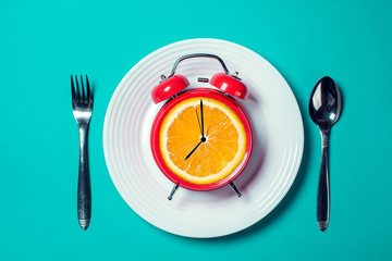 Alarm clock with orange fruit instead of watch dial on the white plate with spoon and fork on the...