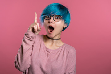 Portrait of young thinking pondering woman with unusual blue hairstyle having idea moment pointing finger up on pink studio background. Smiling happy girl showing eureka gesture.