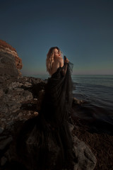 Young woman in long transparent black dress among beach and sea. Wind blows hem of her dress.