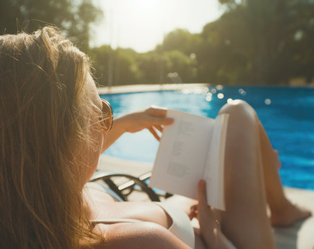 Pretty lady in sunglasses reading book near the pool in the summer.