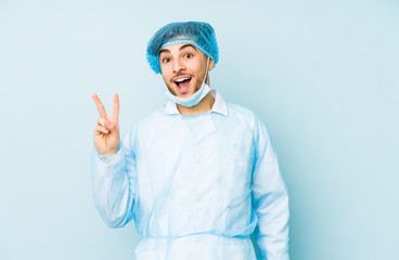 Young arabian surgeon man isolated against on a blue background joyful and carefree showing a peace symbol with fingers.
