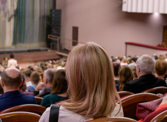 Spectators at the play. Blond woman with shoulder-length hair. The view from the back. The concept of a public event, film screening, presentation, seminar.