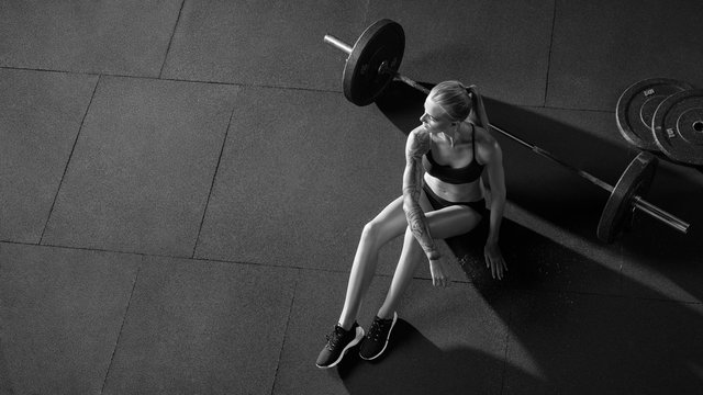Barbell girl Crossfit woman sitting on floor at gym looking into the distance, resting after cross-fit workout with barbell Top view Black and white photo Copy free space.