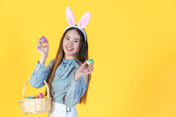 Asian young woman wearing bunny ears showing colorful easter eggs, hold basket filled eggs, toothy smiling happy face. Holidays and people concept isolated on yellow color background, copy space.