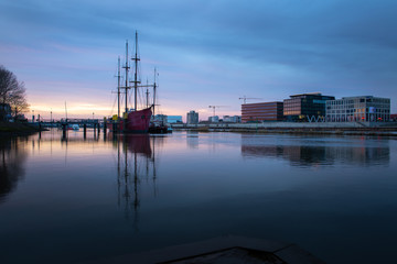 Fototapeta na wymiar long exposure of the Überseestadt in Bremen, Germany with office building, sail boat and perfect reflection on the river weser during blue hour, last light hitting the commercial district