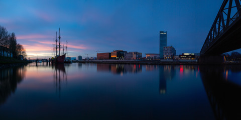 Fototapeta na wymiar long exposure panorama of the Überseestadt in Bremen, Germany with office building, sail boat and perfect reflection on the river weser during blue hour, last light hitting the commercial district
