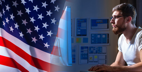A man at a computer creates a website for an American customer. The programmer works against the background of the us flag. Remote work for an American company.