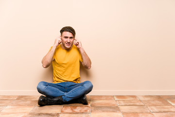 Young caucasian man sitting on the floor isolated covering ears with hands.