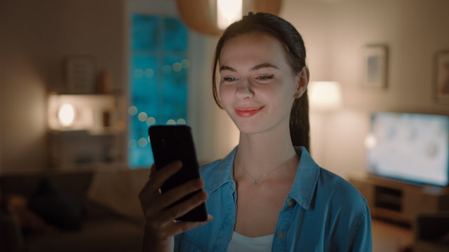 Portrait of Young Beautiful Woman Using Smartphone, Browsing in Internet, Checking Social Networks and Using Smart Home Application while Standing At Home. Cozy Evening.